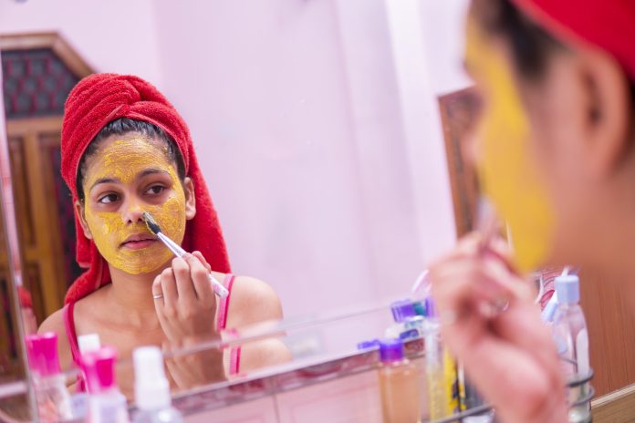 Skin care - Beautiful girl applying Gram flour (Besan) turmeric yellow face mask on face through brush. she is wearing red towel on head and standing front of the mirror at bathroom.