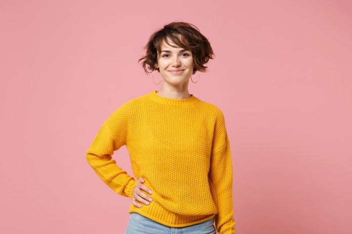 Smiling young brunette woman girl in yellow sweater posing isolated on pastel pink wall background, studio portrait. People sincere emotions lifestyle concept. Mock up copy space. Looking camera