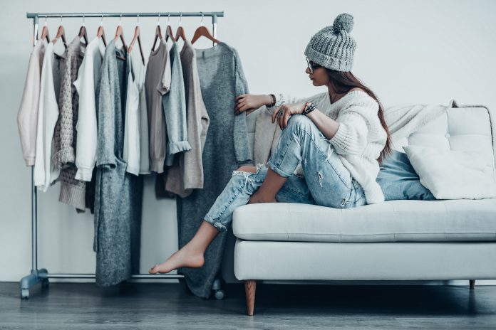 Giovane donna pensierosa in abbigliamento casual che sceglie il vestito mentre è seduta sul divano di casa vicino ai suoi vestiti appesi agli scaffali
