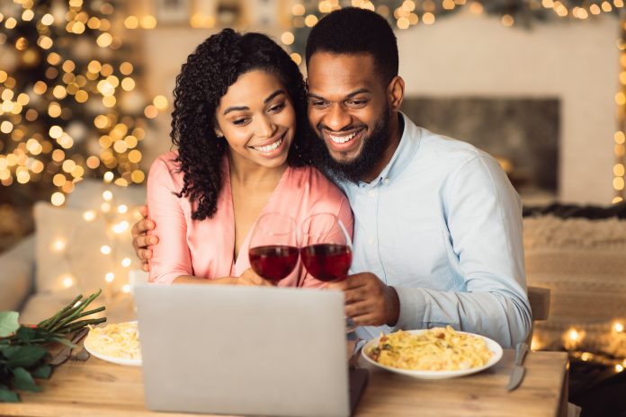 Virtual Party Concept. Smiling african american couple sitting at the table and having online video call with friends or family using laptop, drinking wine and toasting, celebrating together at home