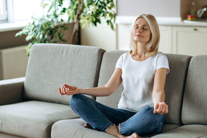 Zufriedene attraktive Frau mittleren Alters sitzt auf dem Sofa zu Hause und chillt. Blonde reifen praktizieren Yoga und Meditation im Lotussitz mit geschlossenen Augen, entlasten Stress, Ruhe Konzept