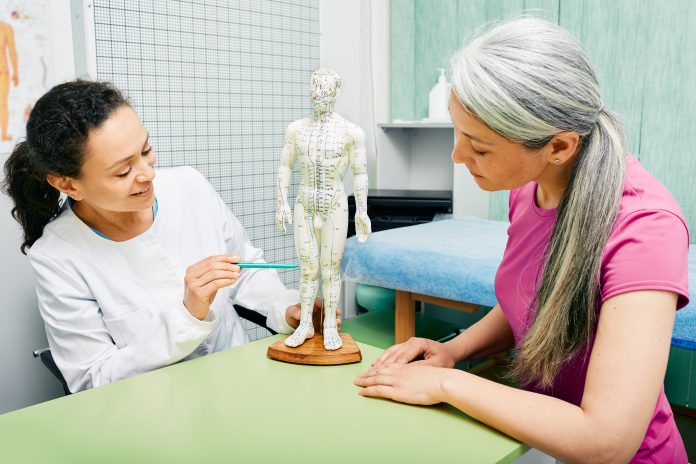Female acupuncturist showing points on acupuncture model of human body to her patient at traditional Chinese medicine centre