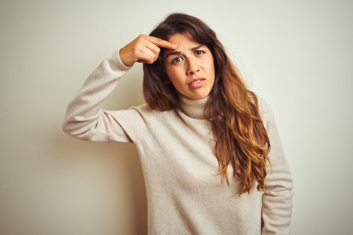 Young beautiful woman wearing winter sweater standing over white isolated background pointing unhappy to pimple on forehead, ugly infection of blackhead. Acne and skin problem